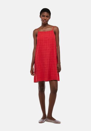 Femme aux cheveux courts portant une robe rouge sans manches à fines bretelles et des chaussures plates en maille claire, debout devant un fond uni.