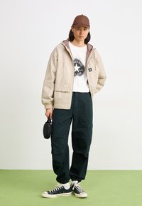 Chaqueta beige con capucha y forro marrón, camiseta gráfica blanca, pantalones cargo verdes oscuros y zapatillas negras. Sosteniendo un pequeño accesorio negro.