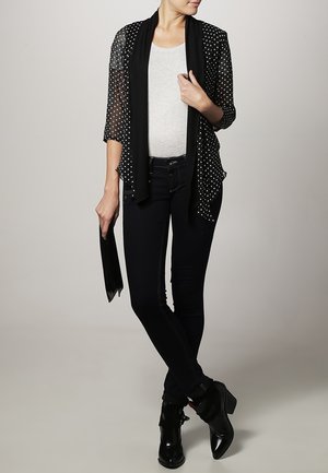Woman wearing black polka dot sheer cardigan over white top, black skinny jeans, and black ankle boots, holding a black clutch.