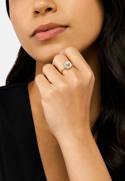 Goldring mit einem runden weißen Edelstein und einem kleinen goldenen Akzentdesign. Die Hand des Models ruht auf dem Kinn und präsentiert die Details des Rings.