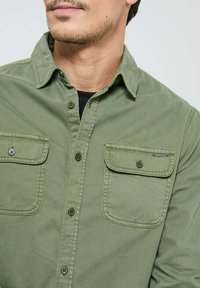 Chemise en boutonnière vert olive en coton, avec un col classique, deux poches poitrine et des boutons contrastés. Texture douce, design décontracté.