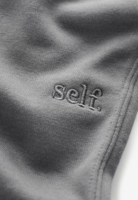 Hallist hallik kangas, millel on pehme tekstuur ja brodéeritud logo, mis ütleb "self." lihtsas ja tagasihoidlikus fondis.