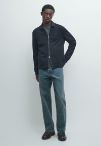 Sort jakke med krave og lynlåslukning foran, kombineret med lyseblå jeans med lige ben og elegante sorte sko. Neutral baggrund.