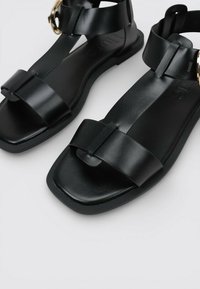 Par de sandalias planas de cuero negro con tiras delanteras anchas, correas en el tobillo y detalles de hebilla dorada sobre un fondo blanco.