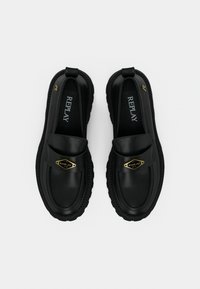 Μαύρα παπούτσια slip-on με λεία υφή δέρματος, διακοσμημένα με χρυσό λογότυπο και χοντρή καουτσούκ σόλα με ανάγλυφες λεπτομέρειες.