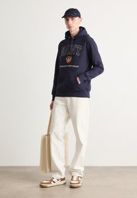 Sudadera navy con el logo "GANT", pantalones crema, zapatillas marrón y blancas, y un bolso beige. Tejido suave, ajuste relajado y estilo casual.