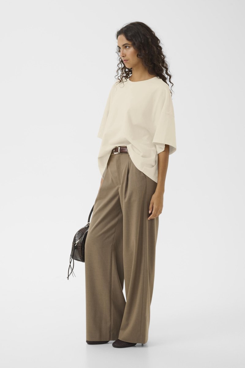 Hellbeige übergroßes T-Shirt, weite braune Hose, dunkle strukturierte Handtasche, kombiniert mit schwarzen strukturierten Schuhen, stehend vor einem schlichten weißen Hintergrund.