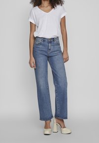 T-shirt blanc léger à col en V associé à un jean large en denim bleu taille haute et des talons compensés crème. Texture douce, coupe décontractée.