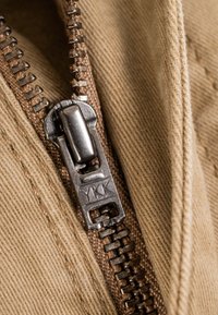 Metal YKK zipper slider partially zipped into beige textile fabric with close-up view of zipper teeth and fabric texture.