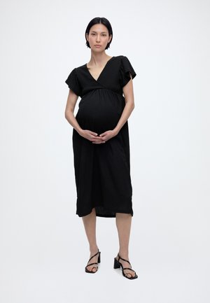Kobieta w ciąży w czarnej sukience do kolan i czarnych sandałach na obcasie, stojąca z rękami delikatnie spoczywającymi na brzuchu na białym tle.