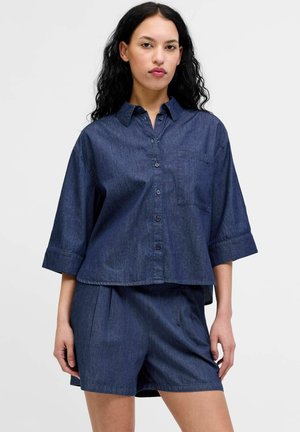 Vrouw met lang zwart haar draagt een losse donkerblauwe blouse met knopen en driekwart mouwen en bijpassende hoge taille shorts, staat rechtop.