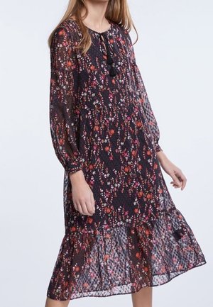 Femme portant une robe florale sombre à manches longues avec un ourlet à volants et un col à nouer avec des pompons, debout devant un fond clair uni.