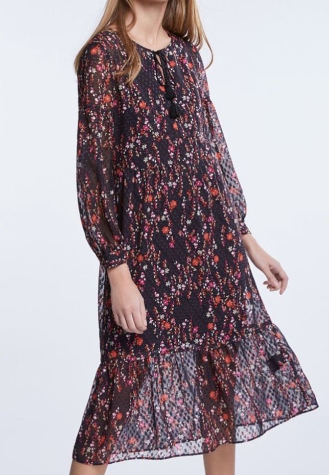 Femme portant une robe florale sombre à manches longues avec un ourlet à volants et un col à nouer avec des pompons, debout devant un fond clair uni.