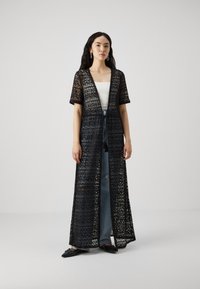 Svart blonder kimono med korte ermer, åpen front, intrikat sikksakkmønster, båret over en hvit topp og blå jeans, som slutter ved anklene.
