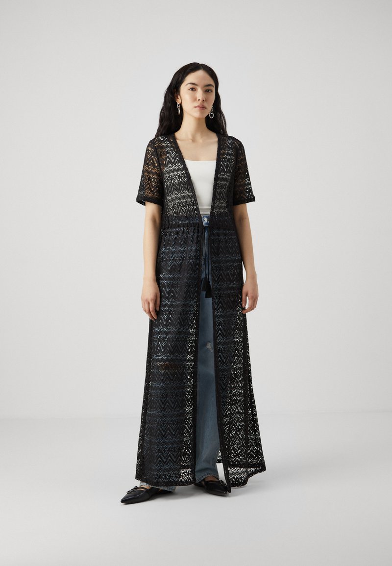 Svart blonder kimono med korte ermer, åpen front, intrikat sikksakkmønster, båret over en hvit topp og blå jeans, som slutter ved anklene.