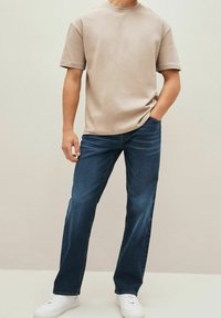 Camiseta de algodón beige, cuello redondo, mangas cortas, combinada con pantalones vaqueros rectos azul marino y zapatillas blancas. Un atuendo simple y casual.