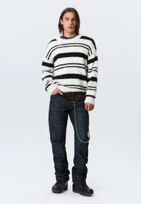 Un jeune se tient debout, portant un pull rayé blanc et noir en peluche, un jean noir avec une chaîne et des bottes noires, le tout sur un fond uni.