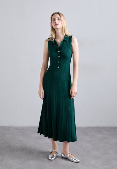 Vrouw in een mouwloze donkergroene gebreide midi-jurk met gouden knopen en zilveren Mary Jane vlakke schoenen, staande op een grijze vloer tegen een grijze muur.