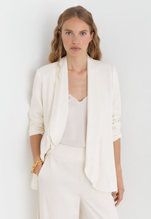 Frau mit langen, welligen Haaren, die einen elfenbeinfarbenen Blazer, ein spitzenbesetztes Top, passende Hosen und ein massives goldenes Armband trägt, steht vor einer weißen Wand.