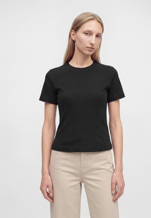 Femme aux cheveux blonds raides portant un haut noir ajusté à manches courtes et un pantalon beige clair, debout devant un fond blanc uni.