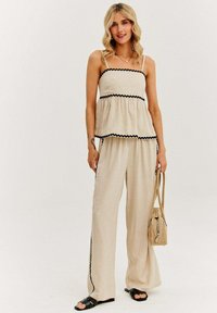 Béžový dvoudílný outfit se zřaseným crop topem a kalhotami širokého střihu, s černými vlnkovými akcenty. Vyrobeno z lehkého materiálu.