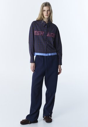 Marineblaue Hose mit weitem Bein, kombiniert mit einem cropped Karohemd, das pinken "NEW AGE"-Text und ein hellblaues Saumdetail aufweist. Weiche Stoffstruktur, legere Passform.