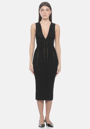 Donna in piedi di fronte che indossa un abito midi nero lavorato a maglia senza maniche con profondo scollo a V e décolleté nere a punta su sfondo bianco.