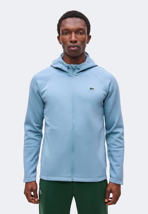 Man draagt een lichtblauwe hoodie met rits en donkergroene broek, staand met een neutrale uitdrukking recht vooruit kijkend tegen een effen achtergrond.