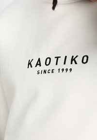 Felpa bianca con un logo nero che riporta "KAOTIKO" e "SINCE 1999" in un font semplice e audace nella zona del petto.