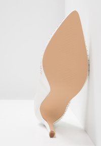 Chaussure blanche à talons hauts avec un bout pointu et une semelle texturée couleur nude. Décorée de petits clous dorés le long des côtés pour un détail supplémentaire.