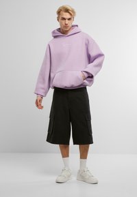 Felpa con cappuccio di colore lilla chiaro, dotata di tasca frontale e logo bianco, abbinata a pantaloni cargo neri con tasche laterali e sneakers bianche.