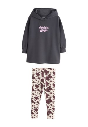 Ciemnoszara bluza z kapturem z napisem „Athletics Studio” w połączeniu z białymi legginsami w brązowy wzór w kształcie plam przypominających krowę.