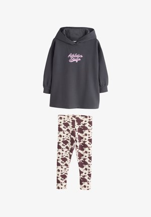 Tamsiai pilkas gobtuvu džemperis su užrašu „Athletics Studio“, derinamas su baltomis tamprėmis, puoštomis rudos karvės raštais.