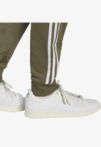 Vita Adidas-sneakers med texturerad överdel, rund tå och ljusfärgad sula. Kombinerade med olivgröna sweatpants med vita ränder.