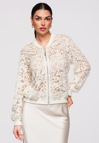 Femme aux cheveux foncés tirés en arrière portant une veste zippée blanche transparente brodée et une jupe en satin crème, posant avec la main sur la hanche.