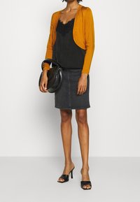 Long-sleeved mustard cardigan over a black lace-trimmed camisole, paired with a black denim skirt and black slide heels. Accessories include a black handbag.