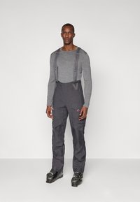 Graues langärmliges Shirt unter dunkelgrauen Schnee-Latzhosen mit verstellbaren Trägern und Taschen, getragen mit schwarzen Skischuhen mit grünen Akzenten.