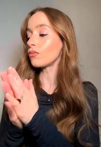 Ung kvinne med langt brunt hår ser til siden, presser hendene sammen, iført en svart topp med lange ermer mot en nøytral bakgrunn.