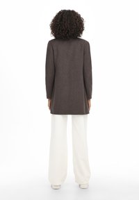 Abrigo marrón de manga larga con una silueta entallada, llevado sobre pantalones blancos de pierna ancha. Textura lisa, diseño minimalista y sin herrajes visibles.