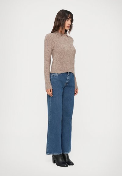 Mujer de pie de lado, vistiendo un suéter de punto beige, jeans de pierna ancha azules y botas negras hasta el tobillo, contra un fondo blanco liso.