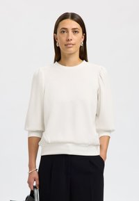 Hvit genser i mykt stoff med rund hals og puffede ermer. Har ribbet kant nederst og på mansjettene. Kombinert med marineblå bukser.