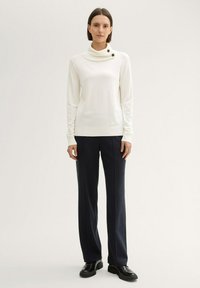 Haut blanc à manches longues côtelé avec un col drapé et deux boutons noirs, associé à un pantalon tailleur bleu marine foncé et des chaussures noires.
