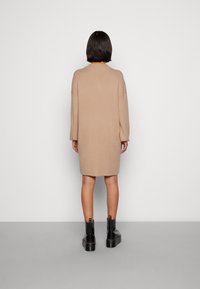 Robe beige en maille avec une coupe décontractée et des manches longues, dotée d'un col rond. Associée à des bottines noires sur une surface grise.