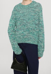 Großer Strickpullover in Teal und Weiß, mit einem runden Ausschnitt und lockerer Passform, kombiniert mit einer kleinen schwarzen Handtasche im Krokodilmuster.