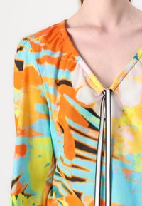Blouse multicolore avec un motif abstrait orange, bleu et jaune. Design en V avec un détail de zip noir et blanc au niveau du col.