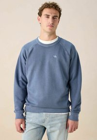 Jeune homme portant un sweat-shirt bleu délavé sur une chemise blanche et un jean bleu clair, se tenant contre un fond beige uni.