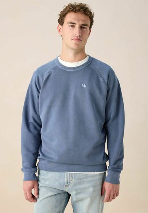 Junger Mann in einem ausgebleichten blauen Sweatshirt über einem weißen Hemd und hellblauen Jeans, der vor einem schlichten beige Hintergrund steht.