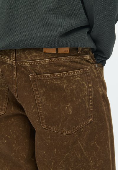 Brown denim pants featuring a distressed finish, visible stitching, and a leather label at the waistband. Back pocket detail is noticeable.