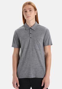 Mladý člověk s dlouhými červenými vlasy, oblečený v šedém polo tričku s krátkým rukávem a náprsní kapsou a v černých kalhotách na jednobarevném pozadí.