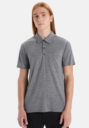 En ung person med långt rött hår som bär en grå kortärmad polo med bröstficka och svarta byxor mot en enfärgad bakgrund.